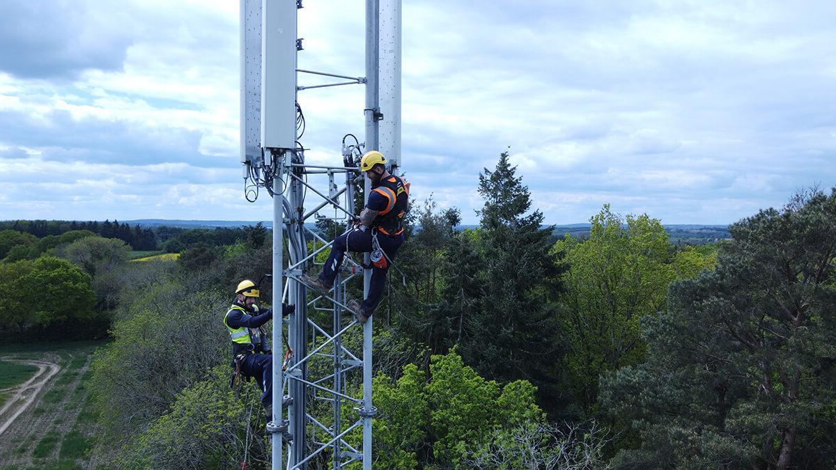 Two Mitie employees at the top of a telecoms tower, with green trees and fields in the background